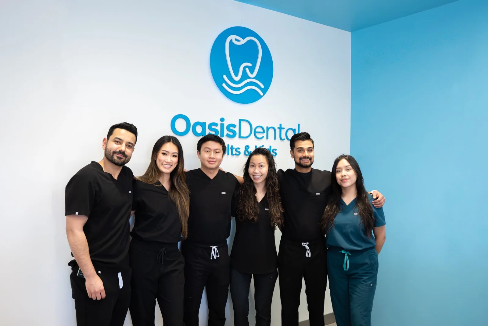 Six Oasis Dental doctors and team members in scrubs standing arm in arm in front of the Oasis Dental logo